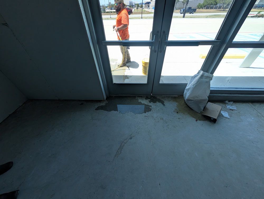 A person in an orange shirt stands outside glass double doors next to a puddle and trash on an unfinished concrete floor.