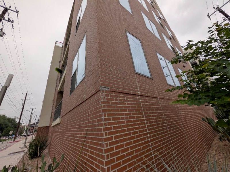 Corner view of a brown brick apartment building beside a sidewalk and trees under an overcast sky