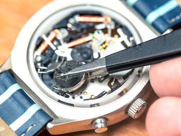A watch being repaired; tweezers holding a small, round battery inside the watch face, blue band visible.