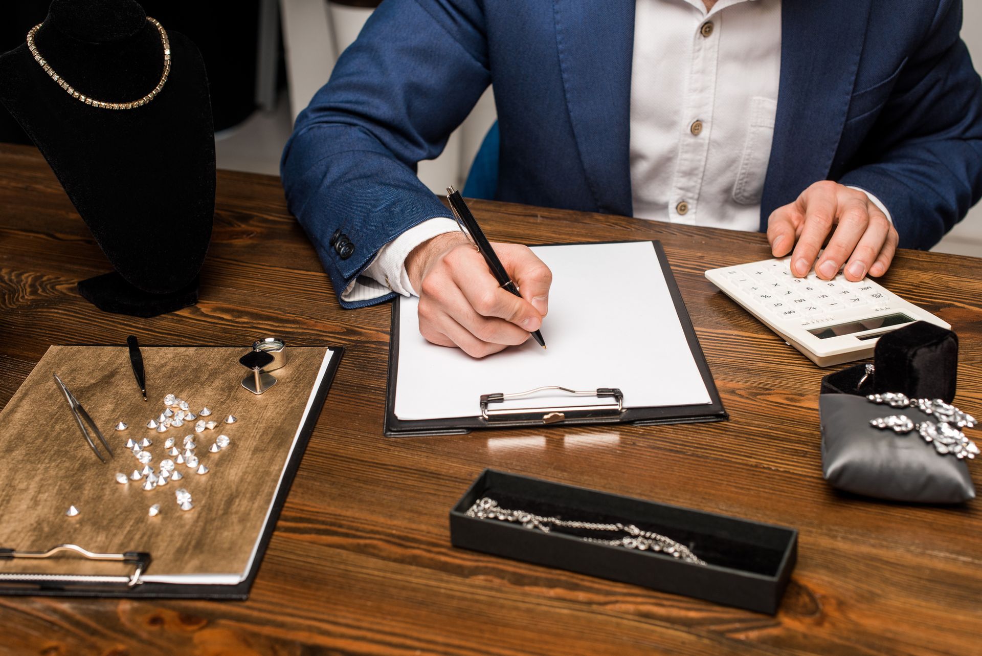 Man in a suit writing on a clipboard, calculating jewelry value on a wooden table.