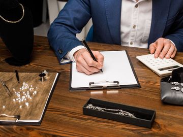 A person in a blue suit, writing on a clipboard at a desk with jewelry and a calculator.