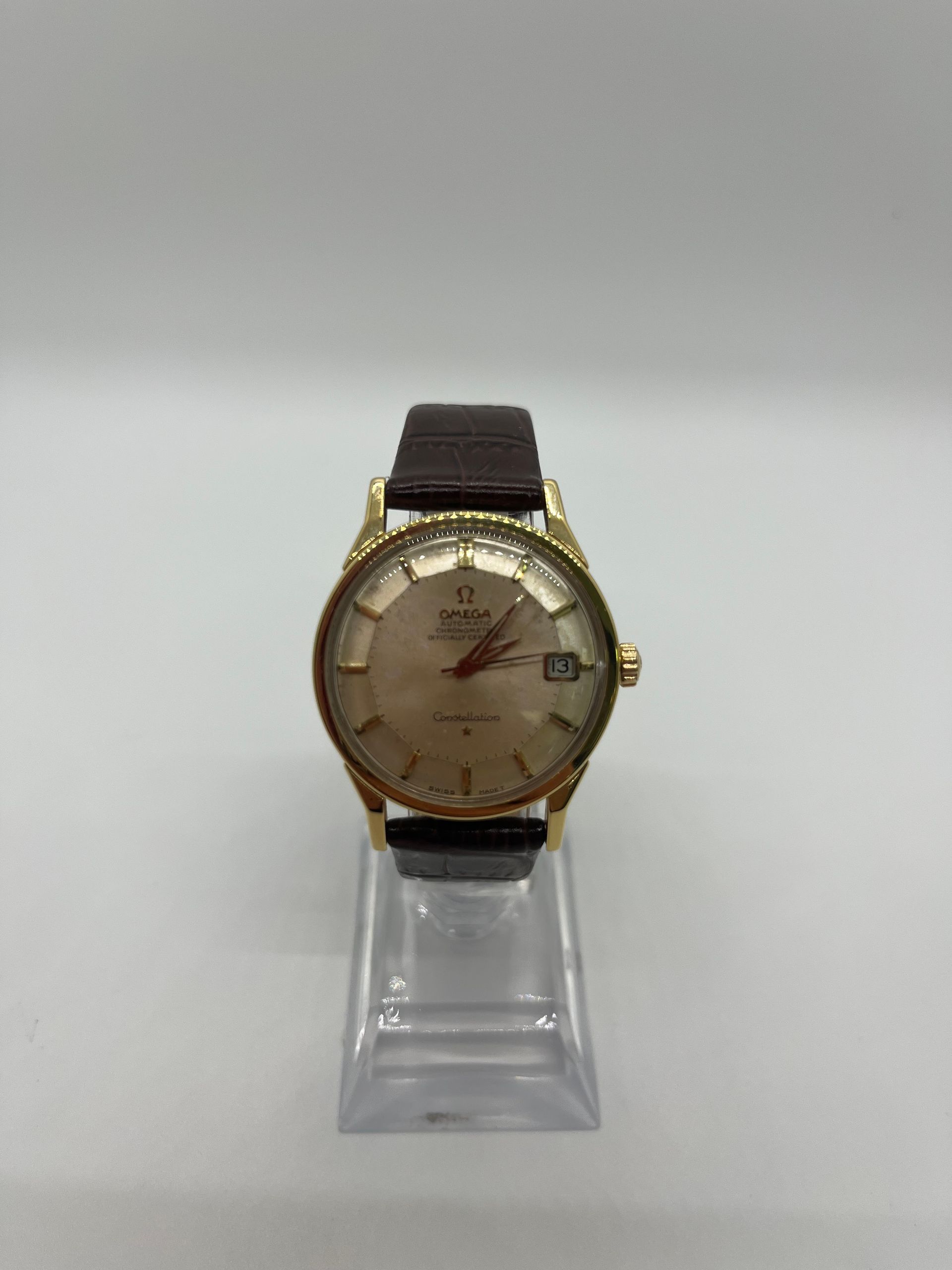 Gold-toned vintage wristwatch on a clear stand; brown leather band, beige face with date window.