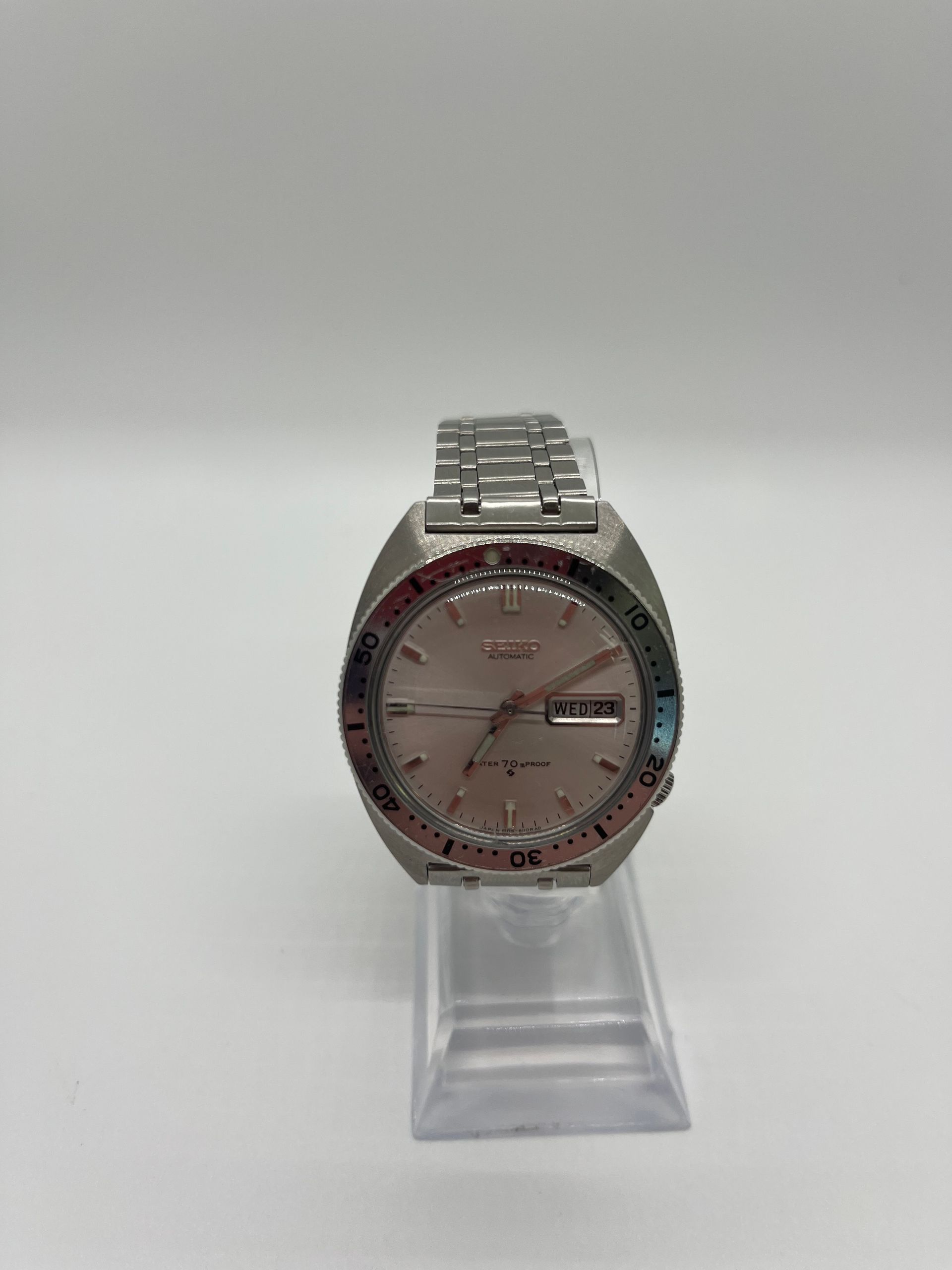 Silver wristwatch with a day/date display on a clear stand, against a white background.