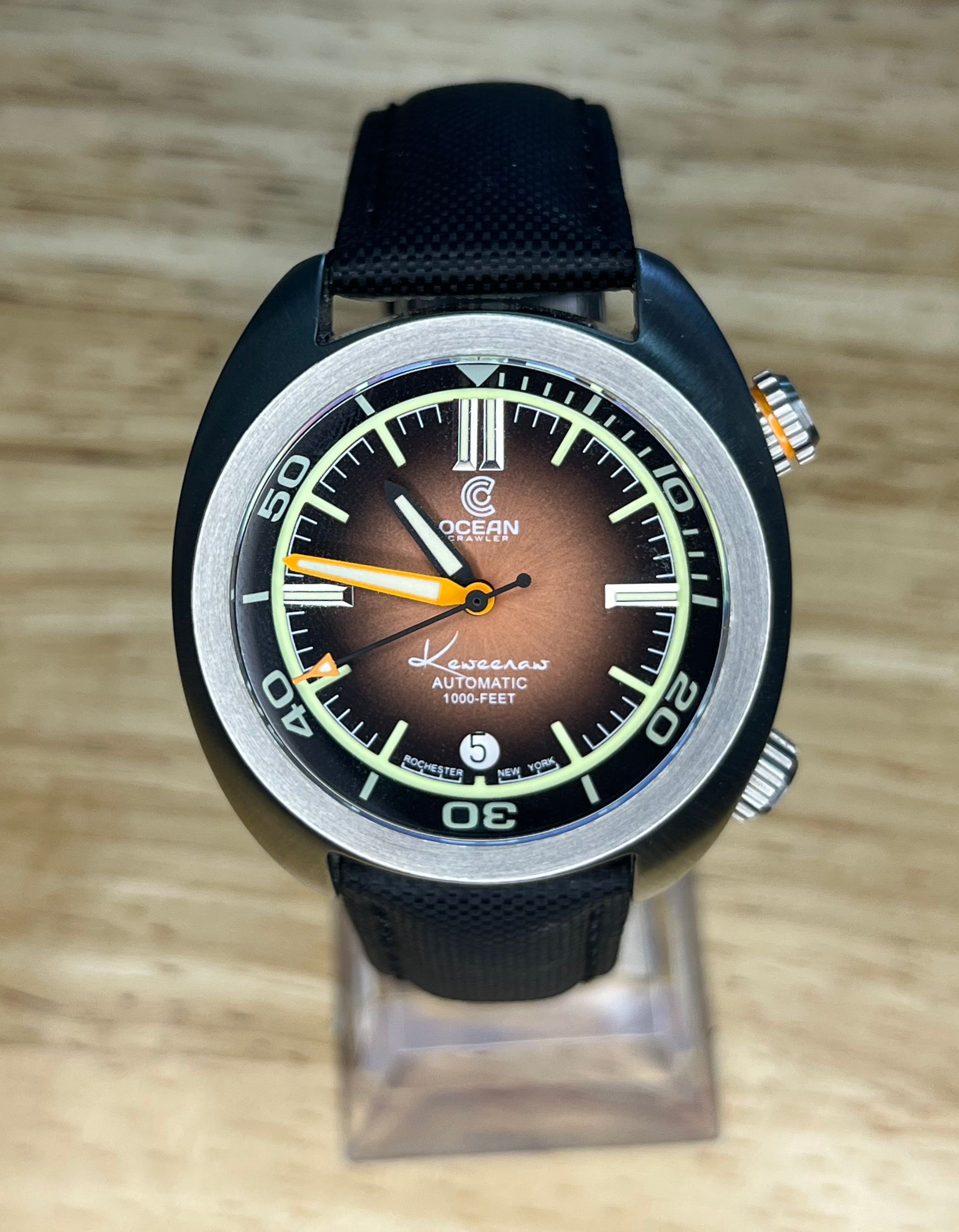 Close-up of a brown-faced, black-cased dive watch with a black strap, resting on a clear stand.