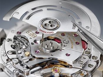 Close-up of a watch mechanism with a metal plate being lifted, revealing intricate internal components.