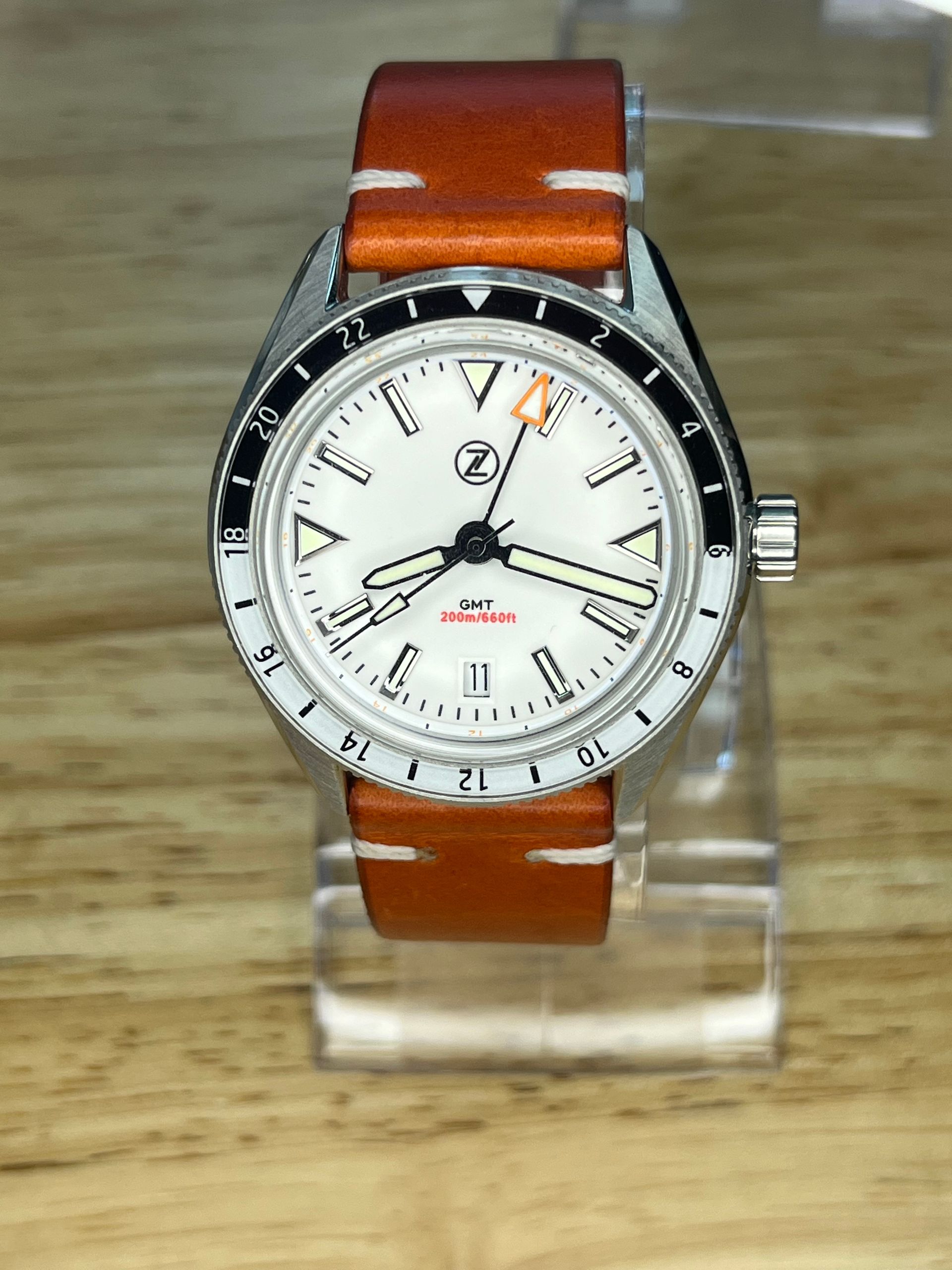 White-faced watch with black bezel and orange leather band, on a clear stand.