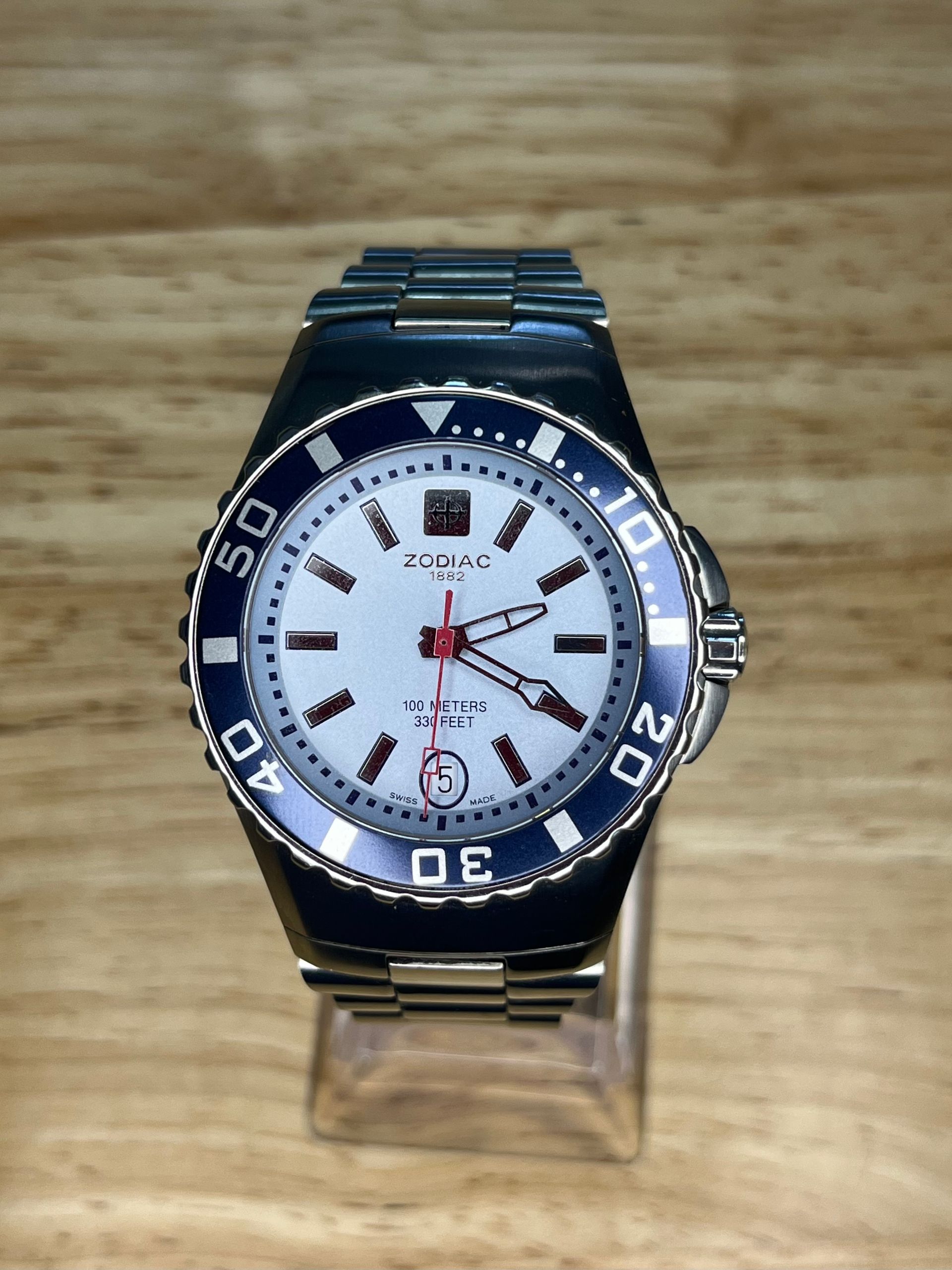 Silver watch with a white face, blue bezel, and silver band on a wooden surface.