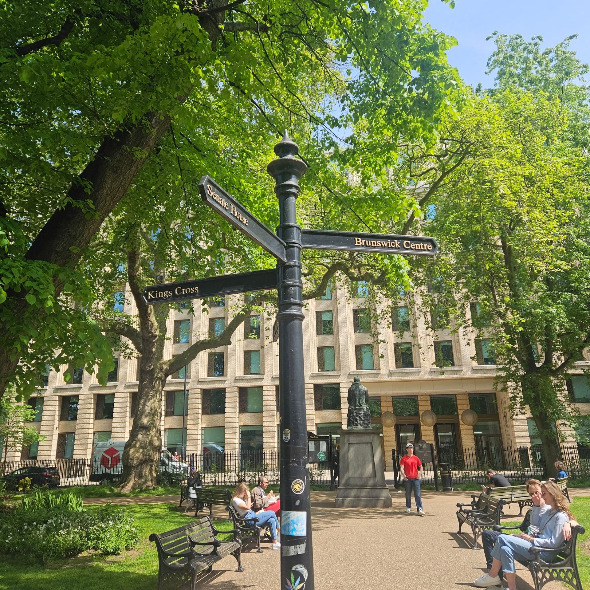 Sunny park with a black signpost, benches, and people sitting near a historic building amid green trees
