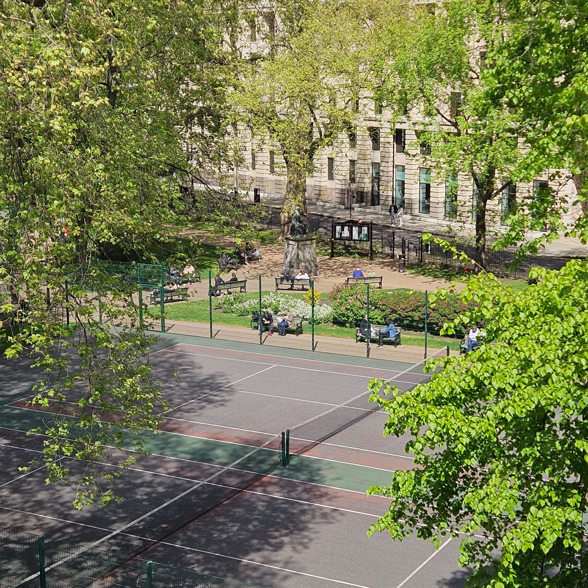 Tree-lined tennis court beside a park, with a few people seated near the fence on a sunny day