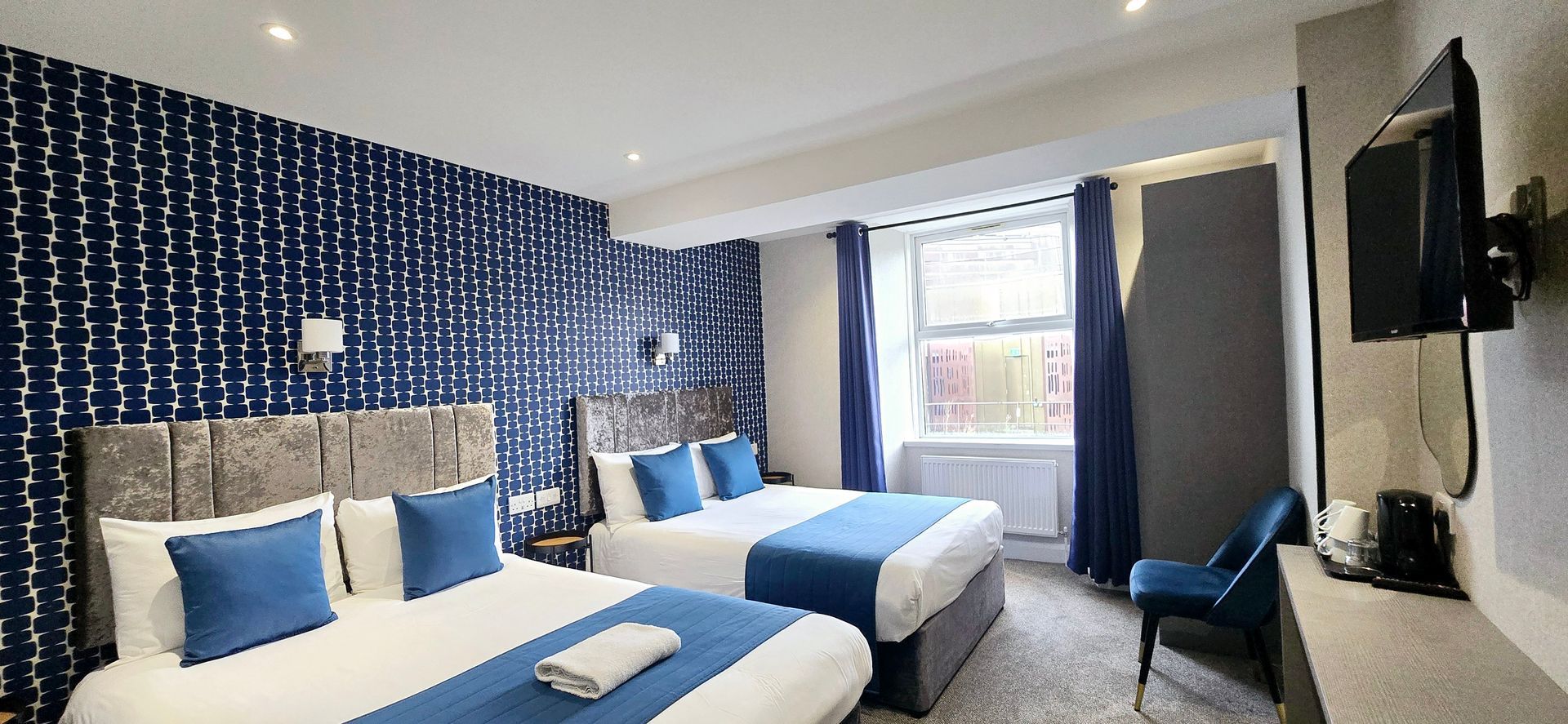 Hotel room with two double beds, patterned blue wallpaper, blue accents, and a window.