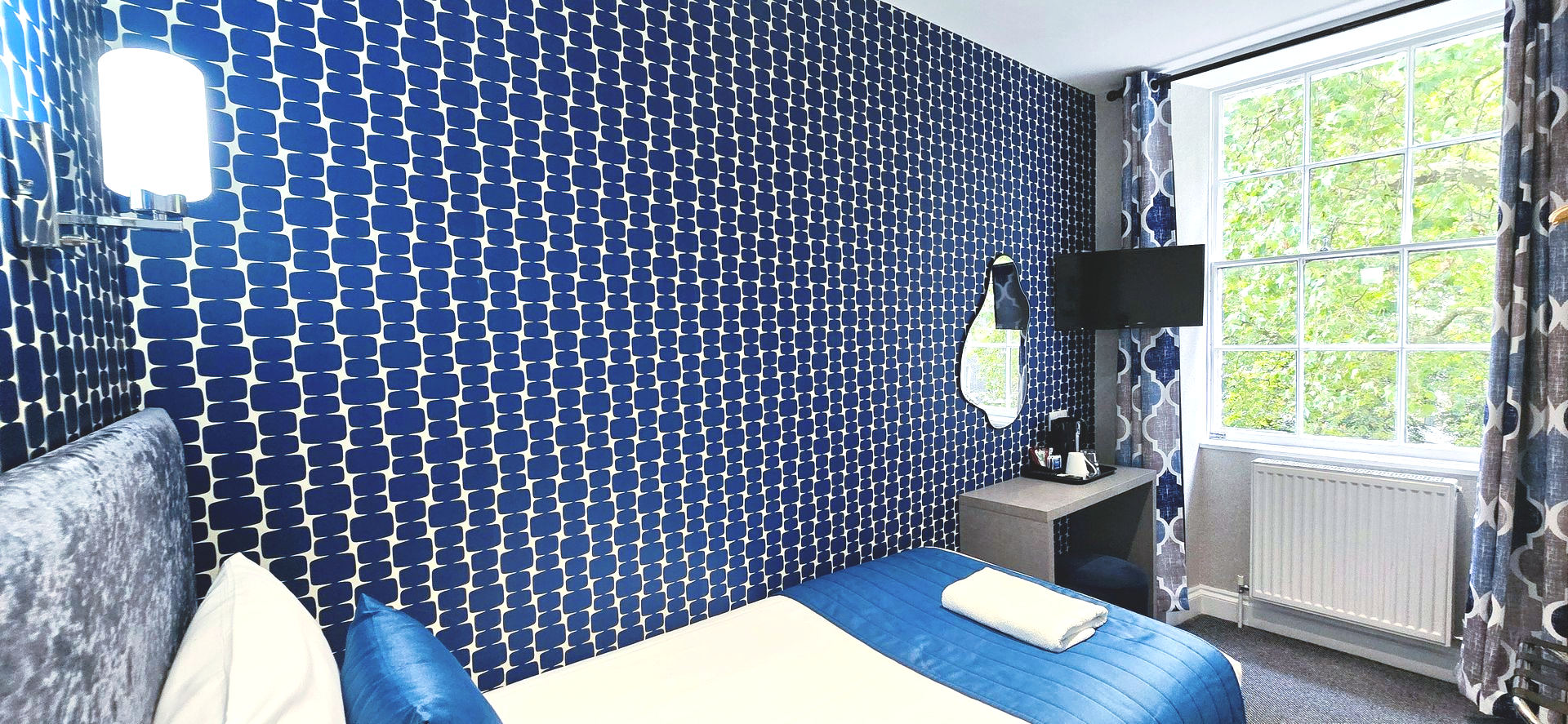 Bedroom with blue patterned wallpaper, a window, and a bed with white sheets and blue accents.