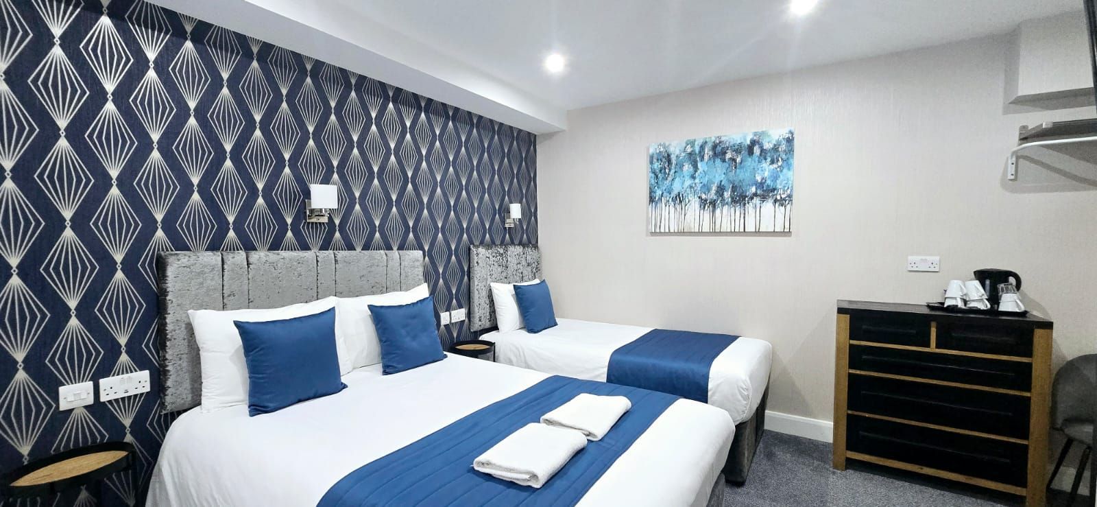 Hotel room with two beds, blue and white decor, patterned wallpaper, and a dresser.