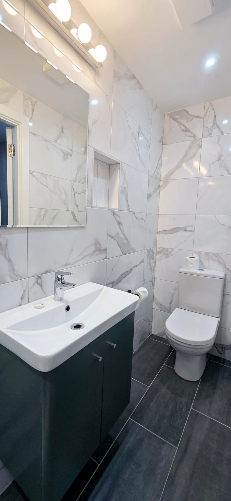 A modern bathroom with marble walls, dark floor tiles, a sink, toilet, and a mirror.