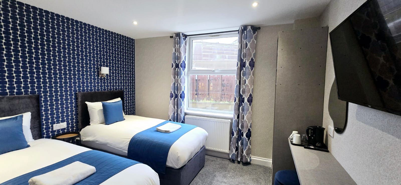 A hotel room with two twin beds, blue accents, patterned wallpaper, and a window.