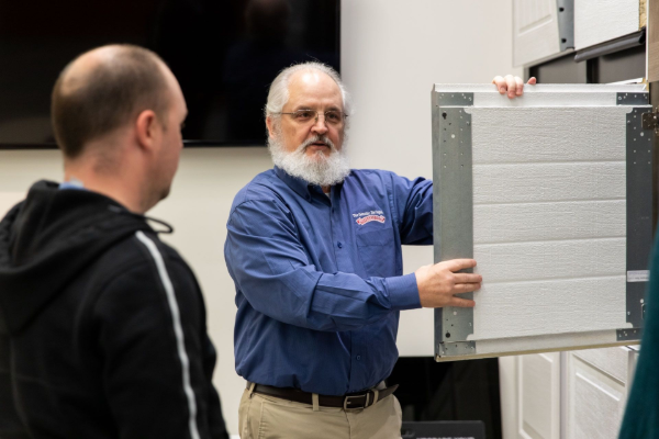 Man in blue shirt demonstrating a large gray appliance panel to another person in a workshop