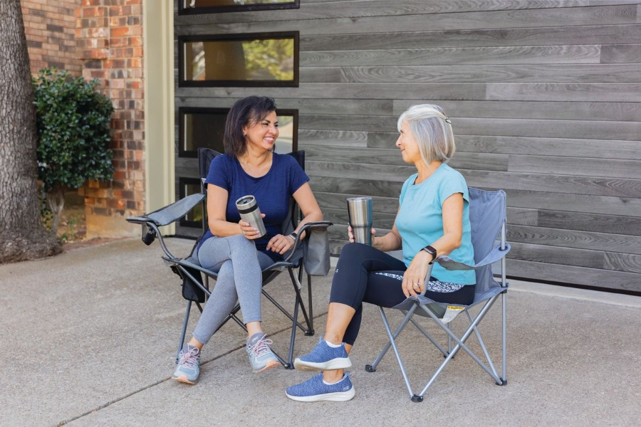 Two women sit in folding chairs chatting and holding mugs outside a wooden building.