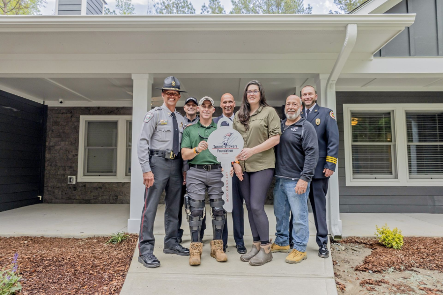 Group standing on a front porch, smiling, with a house and garden in the background.