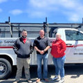 Three people standing beside a white service truck on a sunny street.