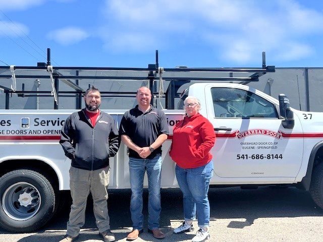 Three people standing beside a white service truck on a sunny street.