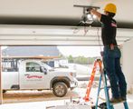 Worker on ladder repairing a garage door opener above a white pickup truck in a snowy driveway