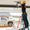 Worker on ladder installing a garage door opener inside a garage, with a work truck outside.