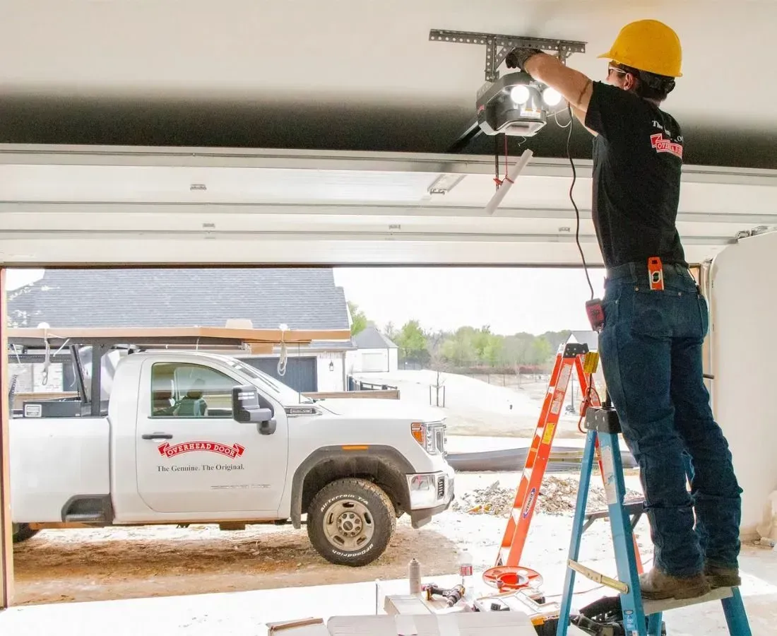 Worker on ladder installing a garage door opener inside a garage, with a work truck outside.