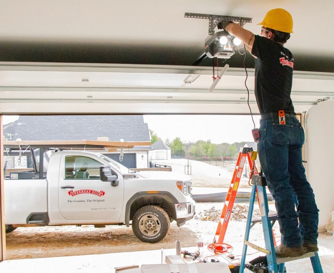 Worker on ladder repairing a garage door opener above a white pickup truck in a snowy driveway