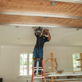 Worker on a red ladder installing wiring in an unfinished room with exposed ceiling beams.