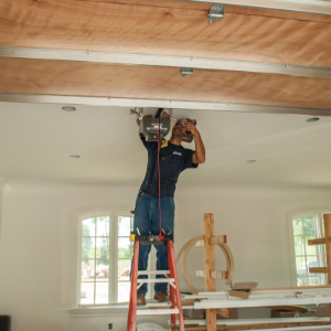 Worker on a red ladder installing wiring in an unfinished room with exposed ceiling beams.