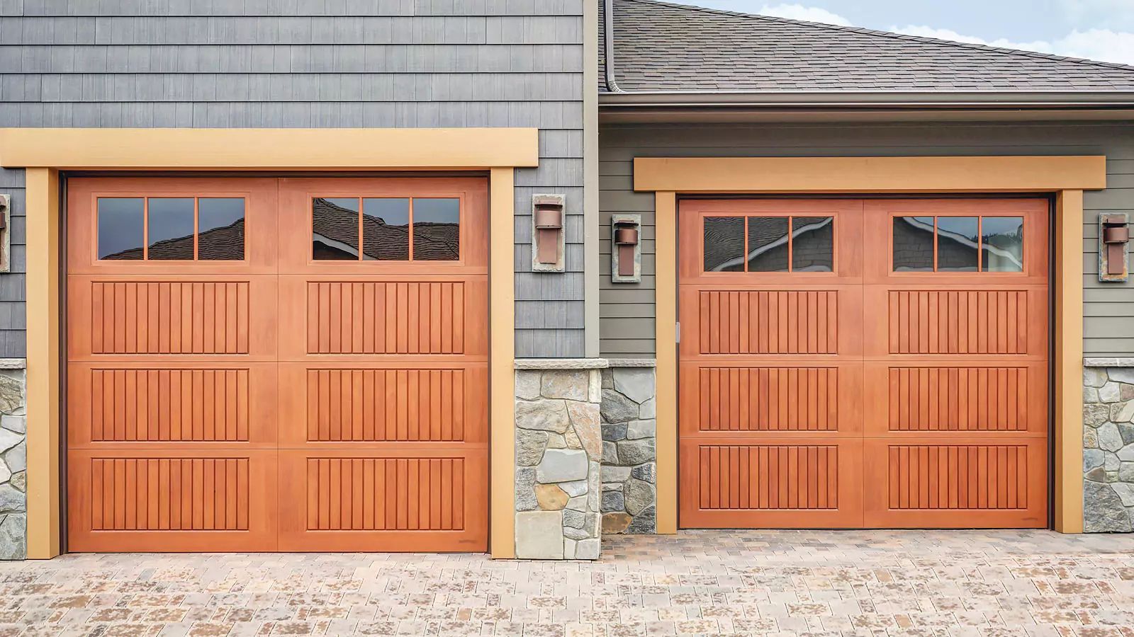 Two wooden garage doors with small windows in a gray stone and siding house facade