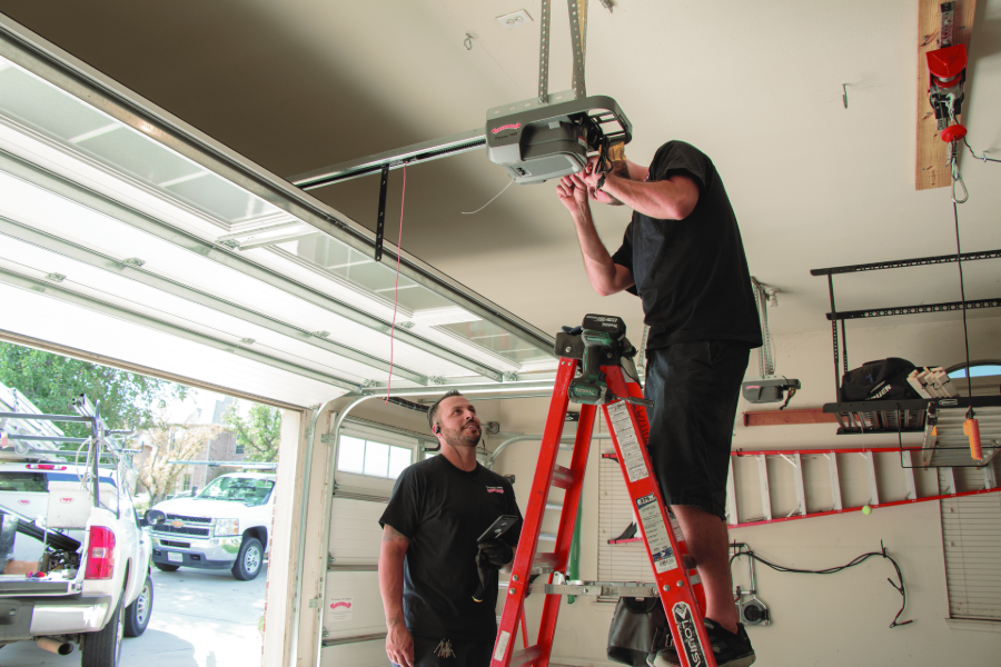 Two workers installing a garage door opener, one on a red ladder under an open garage door.