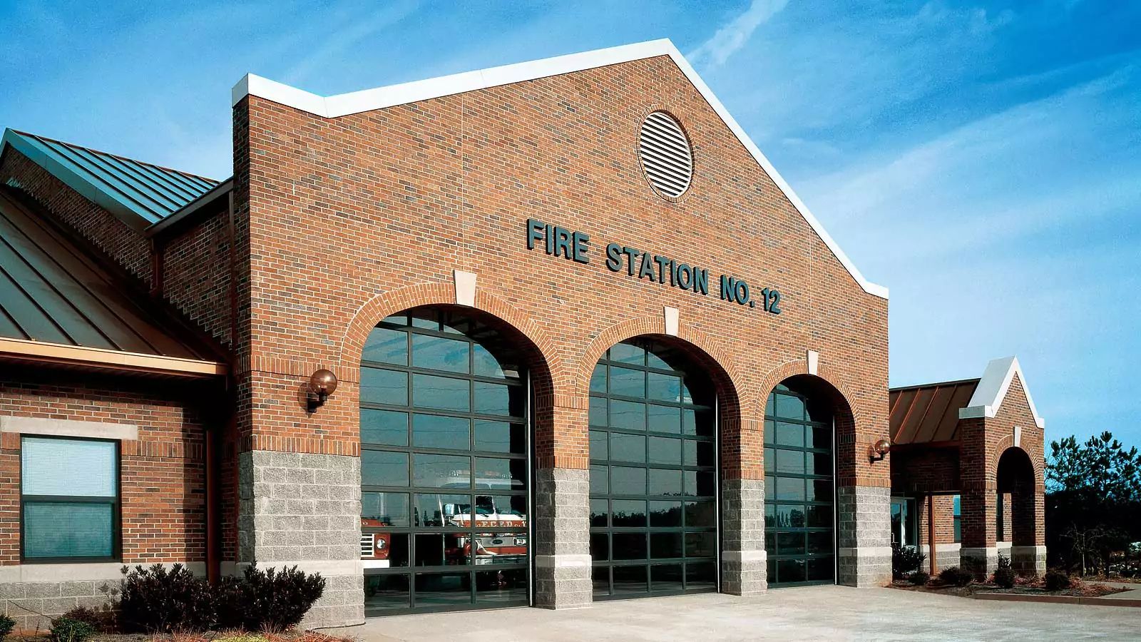 Brick fire station building with arched glass entrances and a metal roof under a blue sky