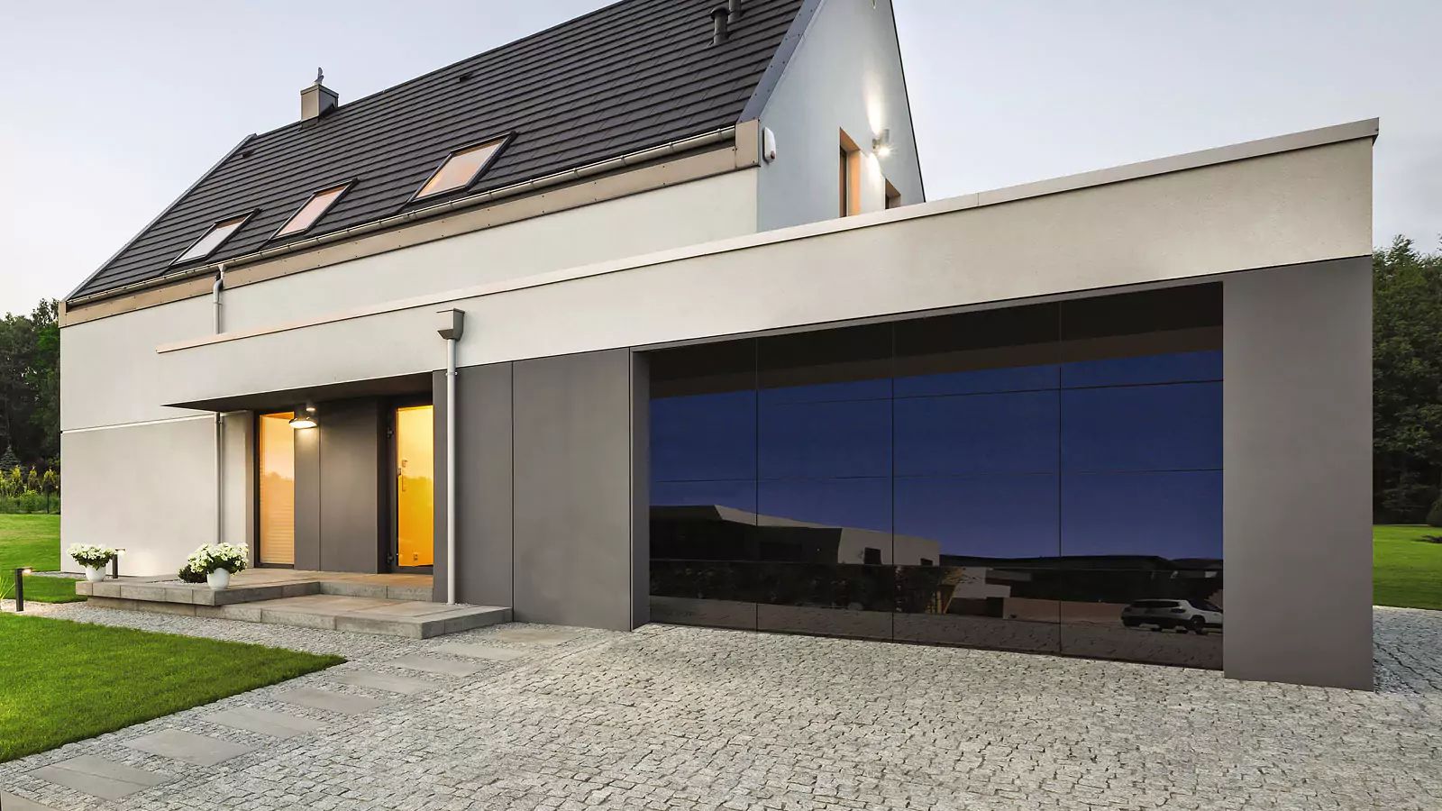 Modern house exterior with a gray facade, dark roof, and large reflective blue windows beside a gravel driveway