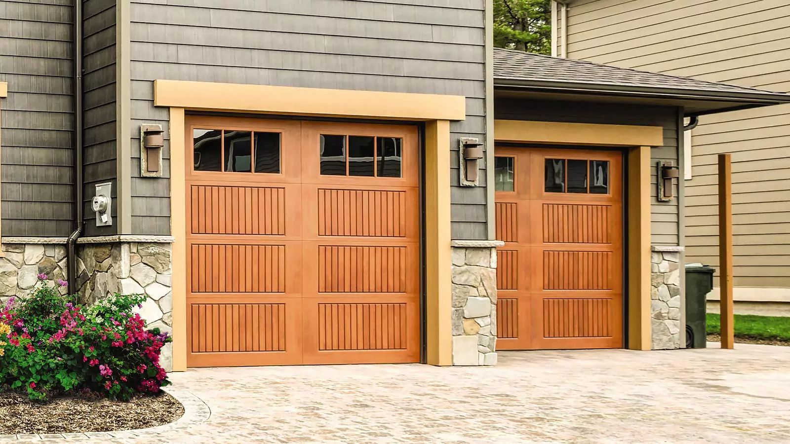 Two orange wooden garage doors on a gray house exterior with a stone driveway and flower bed.
