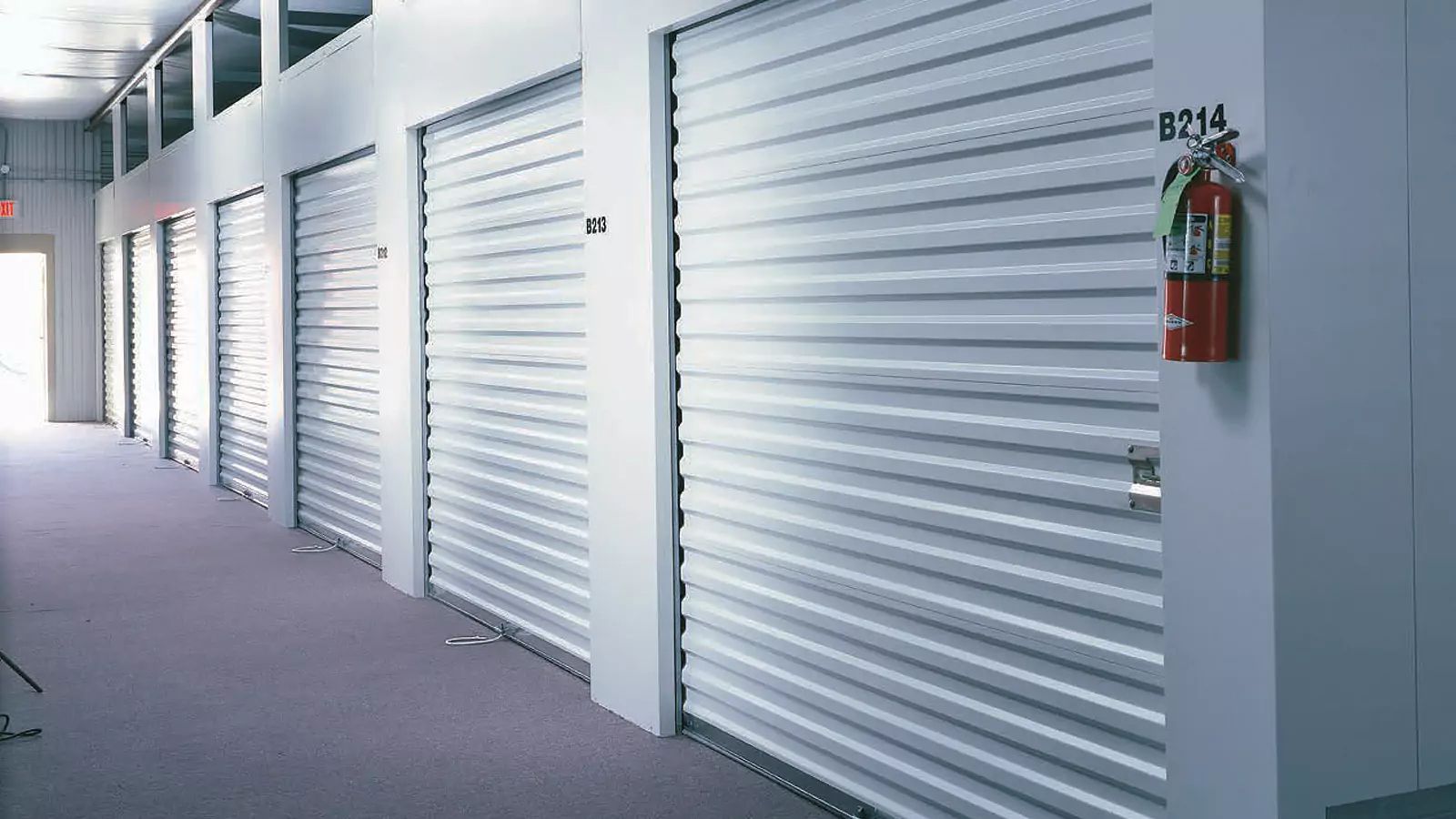 Row of white roll-up storage doors in a clean hallway, with a red fire extinguisher on the wall.