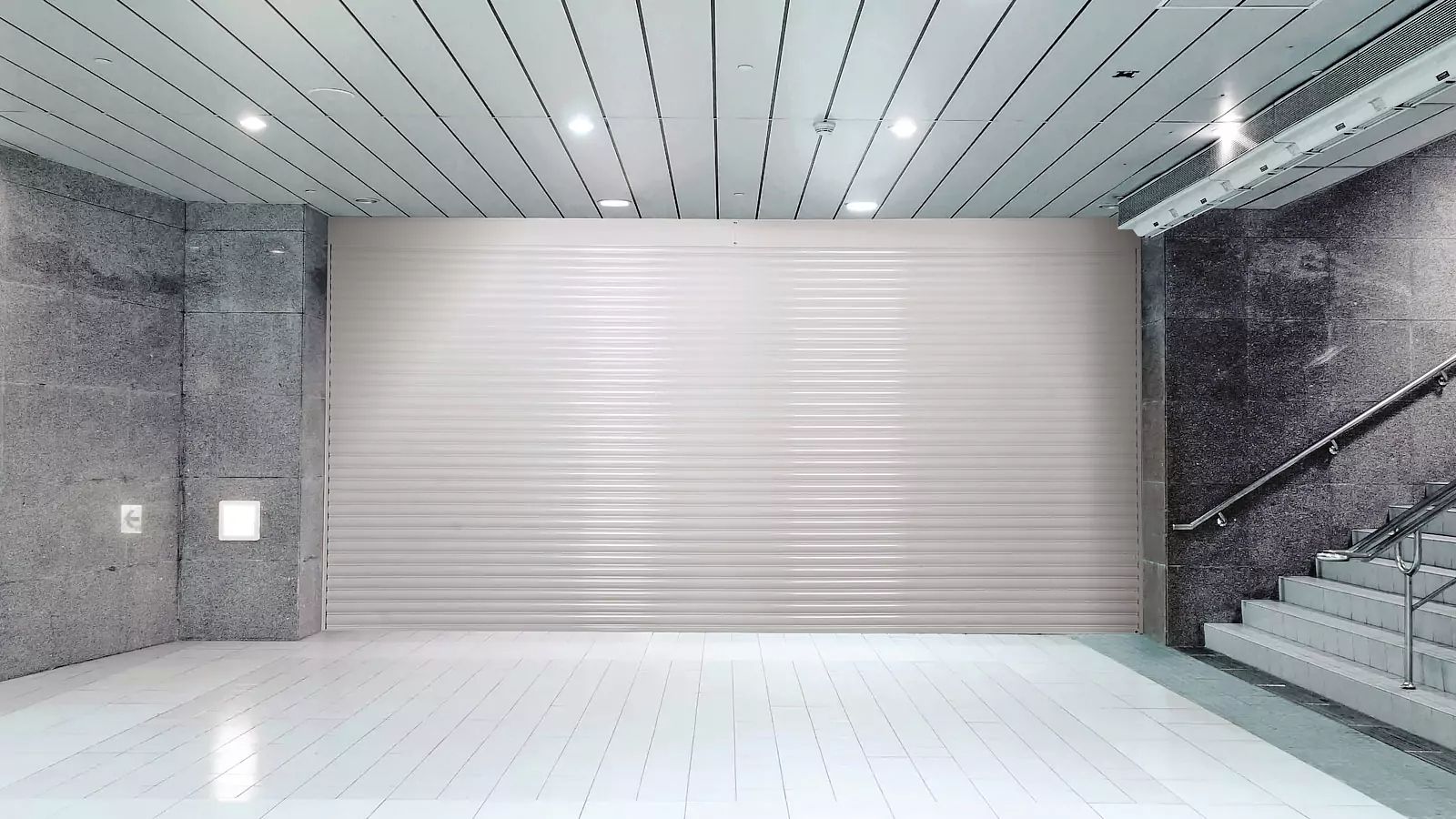 Empty modern lobby with a closed metal shutter door, white tiled floor, and stairs on the right.