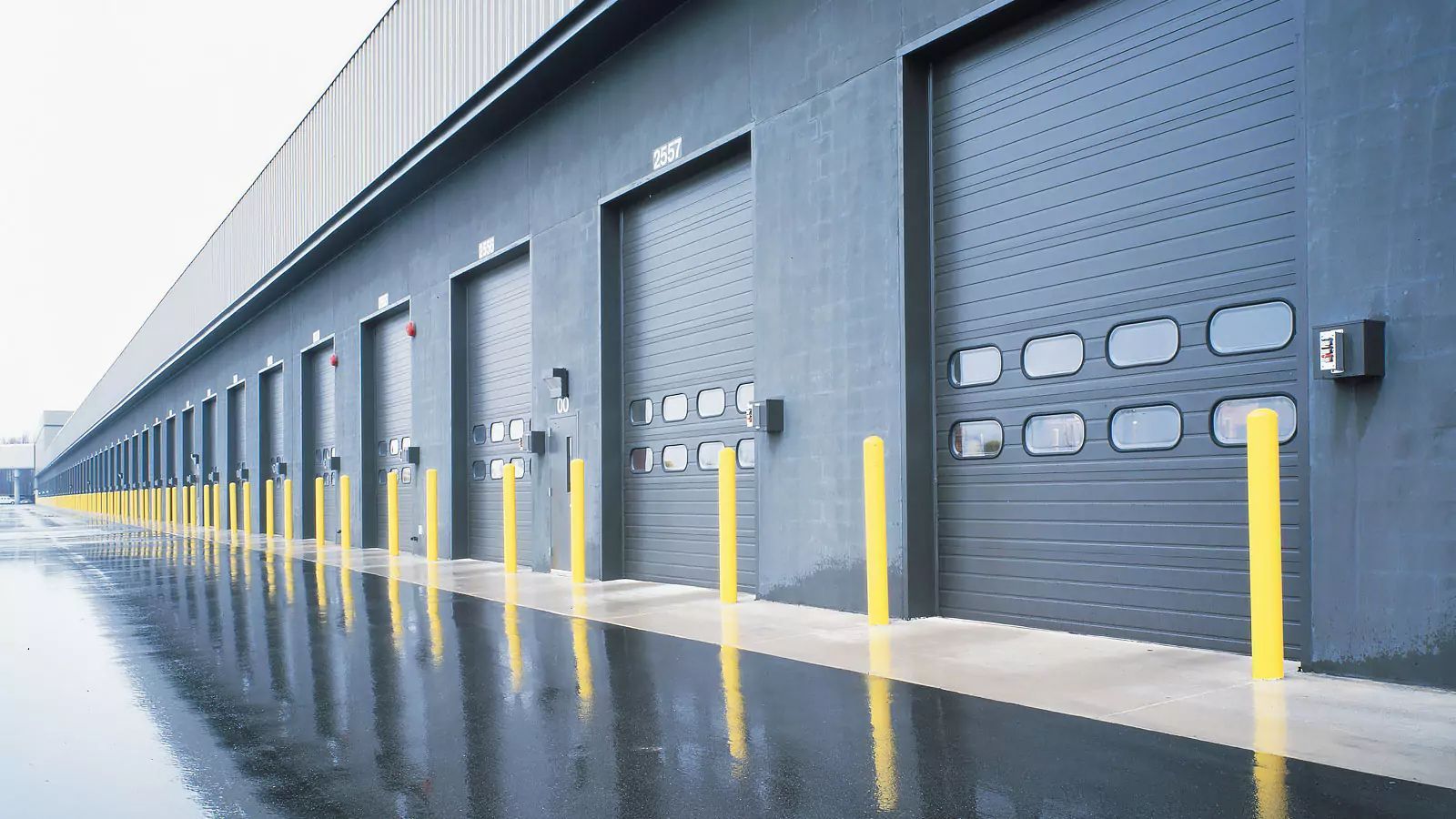 Row of gray warehouse loading docks with yellow safety bollards on a wet concrete surface