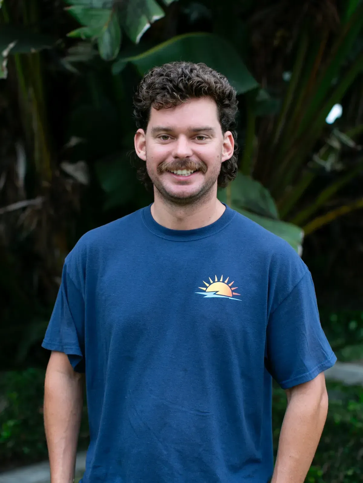 Smiling person in a blue T-shirt standing outdoors in front of leafy greenery
