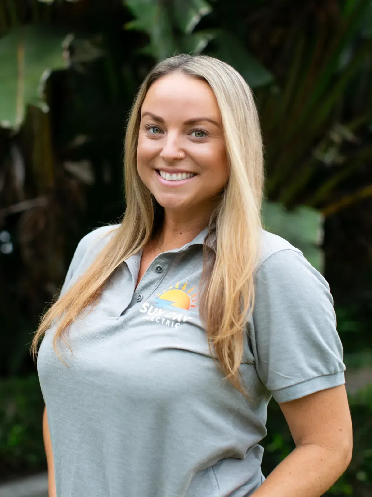 Smiling woman in a gray polo shirt standing outdoors against lush green foliage