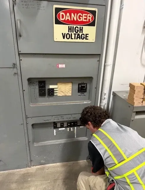 Worker crouches at a high-voltage electrical panel marked “Danger High Voltage”