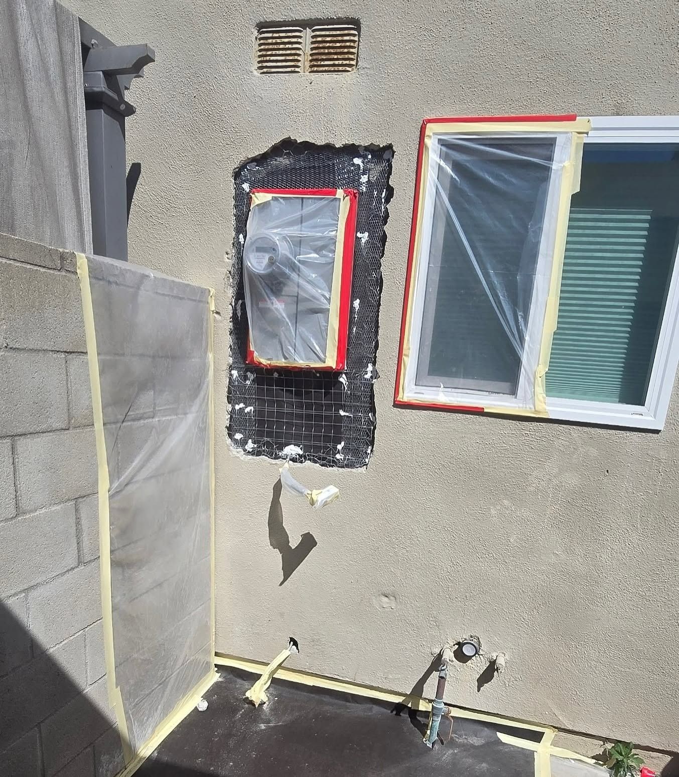 Exterior wall with two taped-over windows, one surrounded by black plastic sheeting and red tape