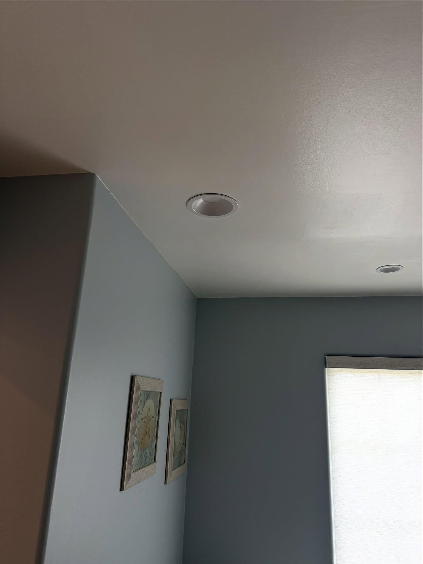 Corner of a light gray room with a white ceiling, recessed light, wall art, and a bright window with blinds