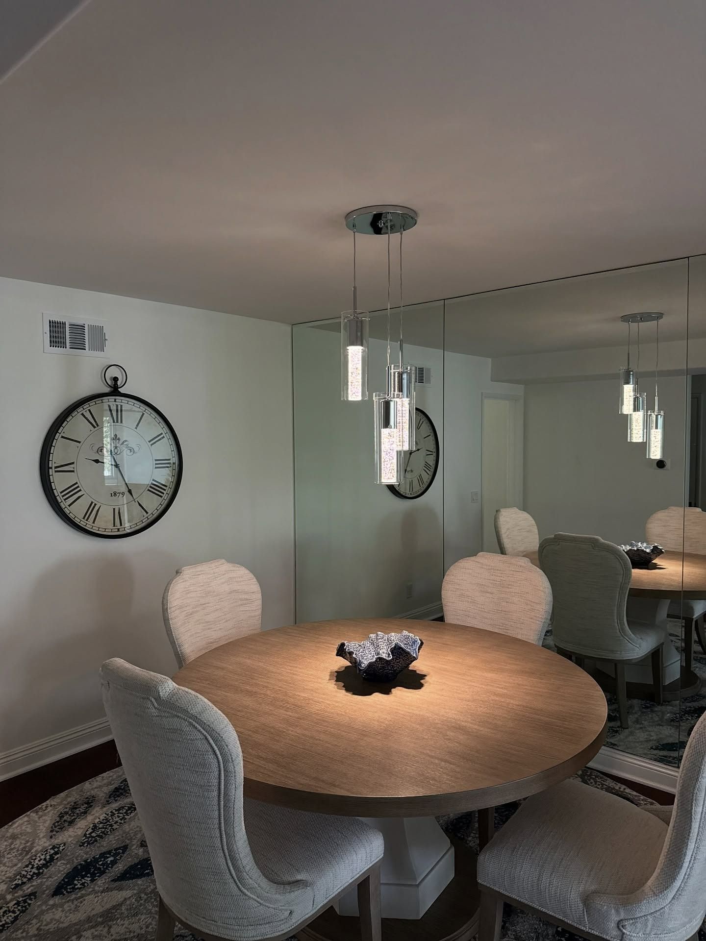 Round dining table with four chairs in a mirrored room, with a large wall clock and hanging pendant lights