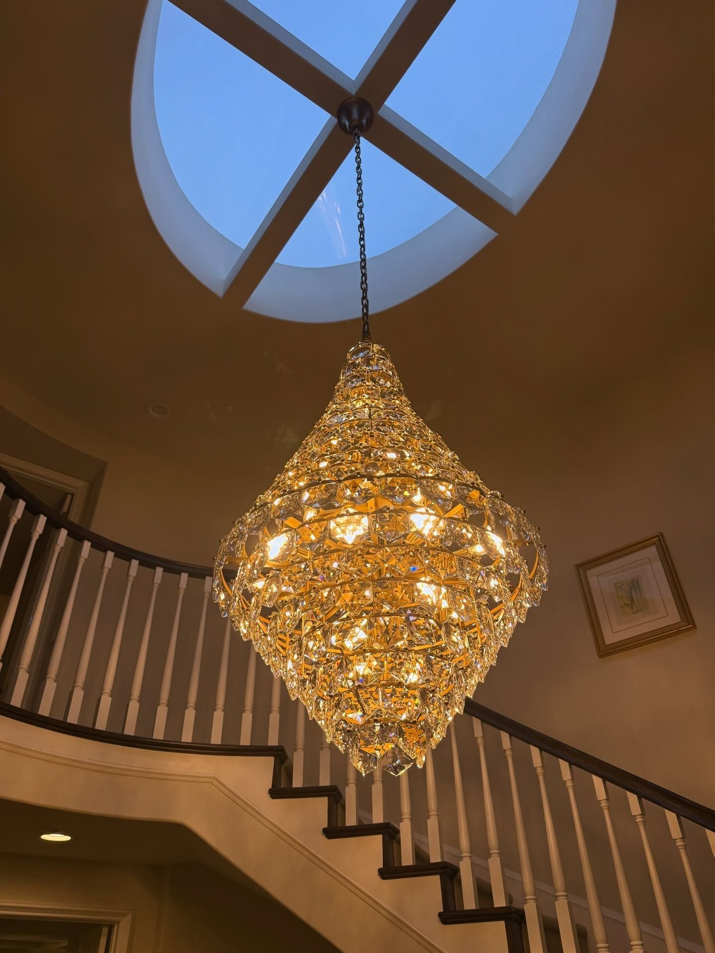 Grand chandelier hanging beneath a skylight above a curved staircase in a warm-toned foyer