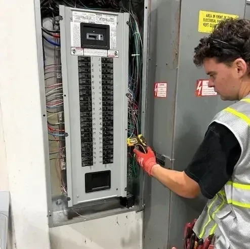 Technician in safety vest wiring an open electrical breaker panel in a utility room