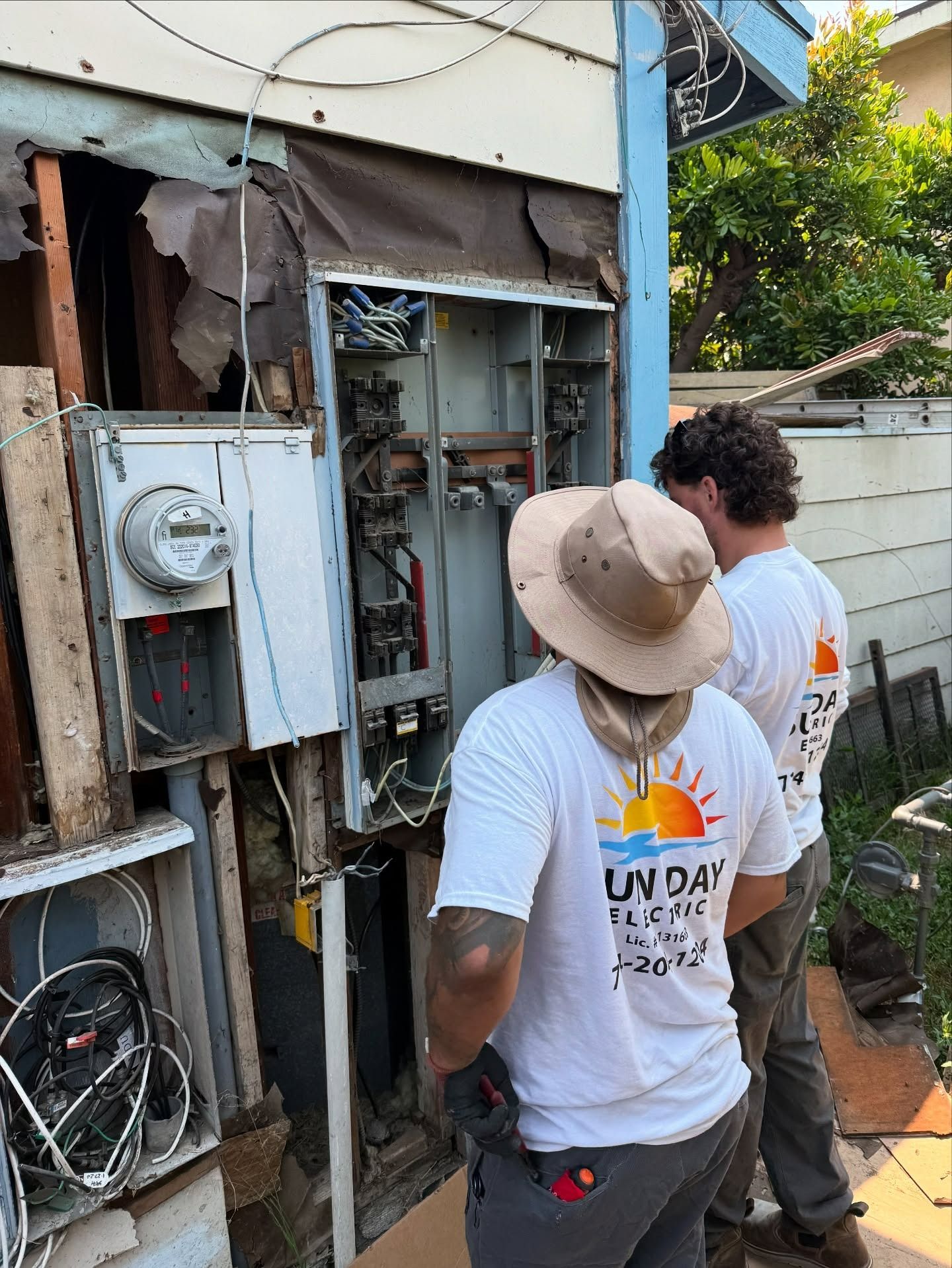 Two workers inspect exposed electrical wiring and meters on a damaged wall outdoors.
