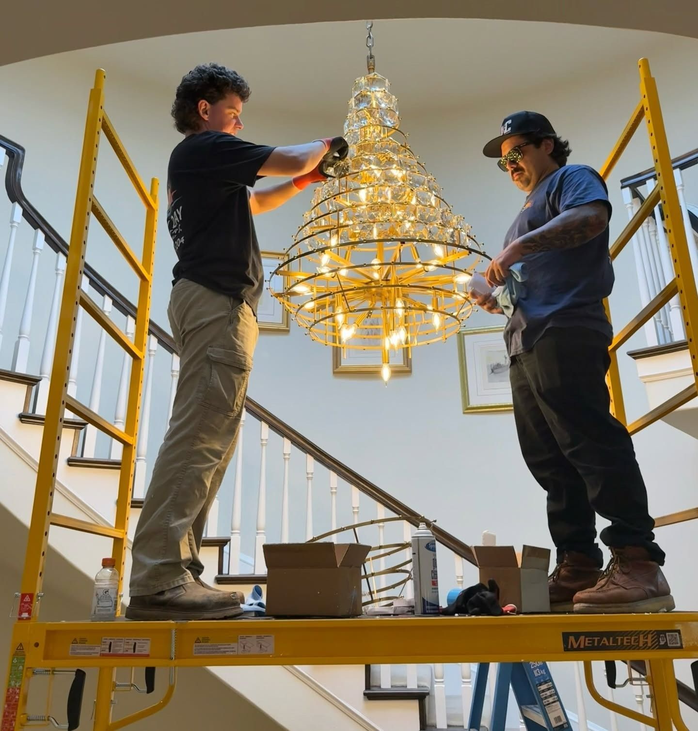 Two workers on a ladder platform install a gold chandelier in a stairwell.