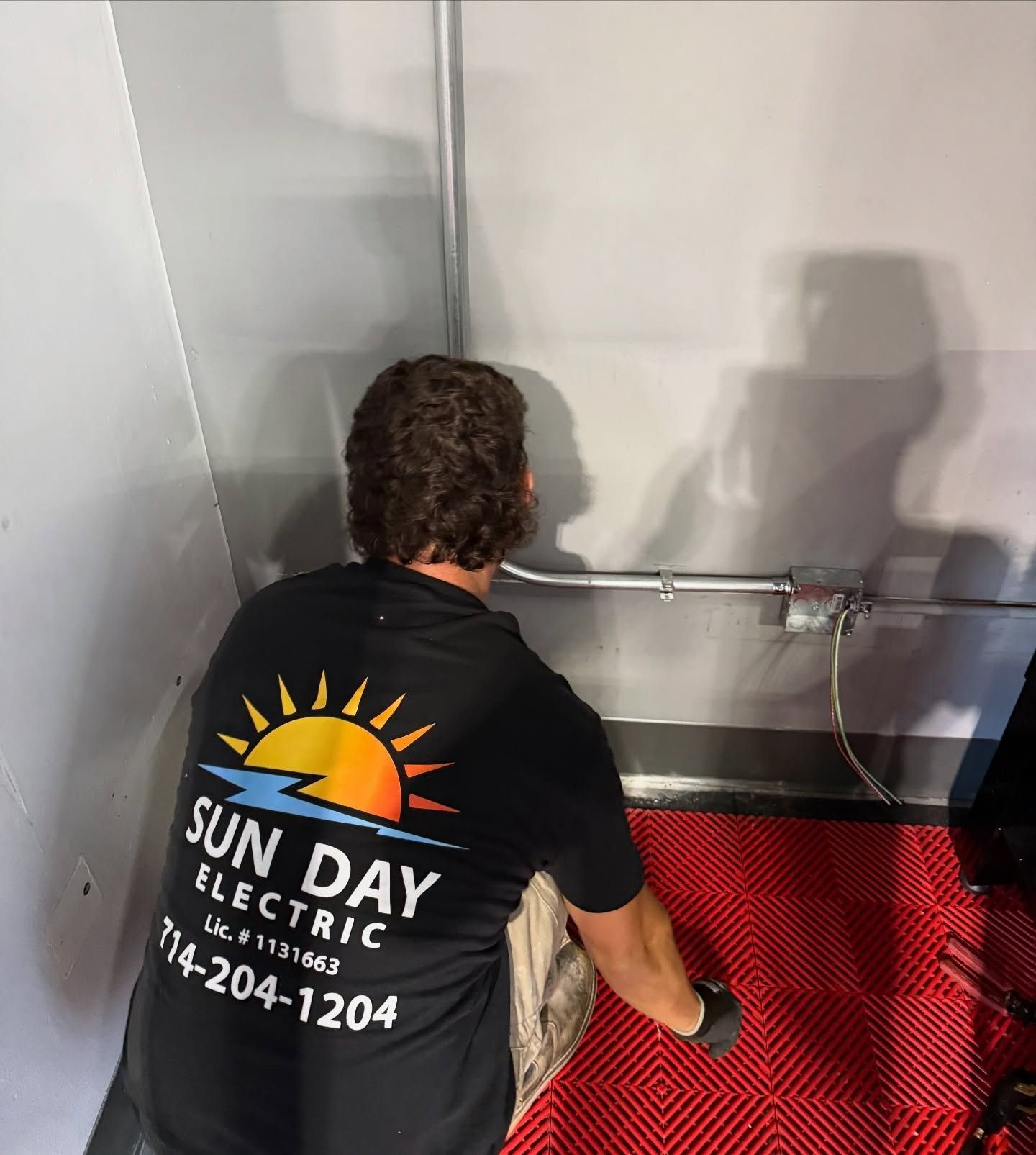 Technician in black Sun Day Electric shirt working on red mat in a corner by a wall-mounted pipe