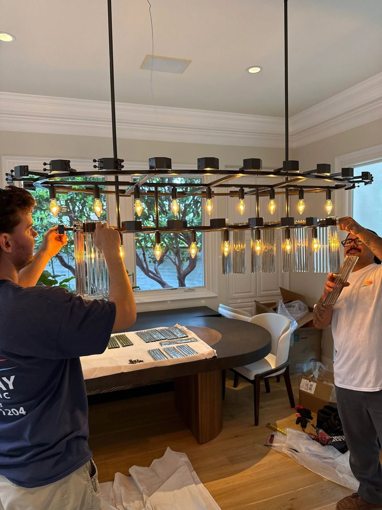 Two people hanging string lights in a dining room with a table and window bench