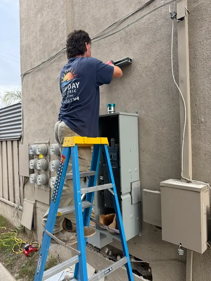 Worker on a blue ladder cleaning an exterior wall beside utility boxes with a pressure washer.